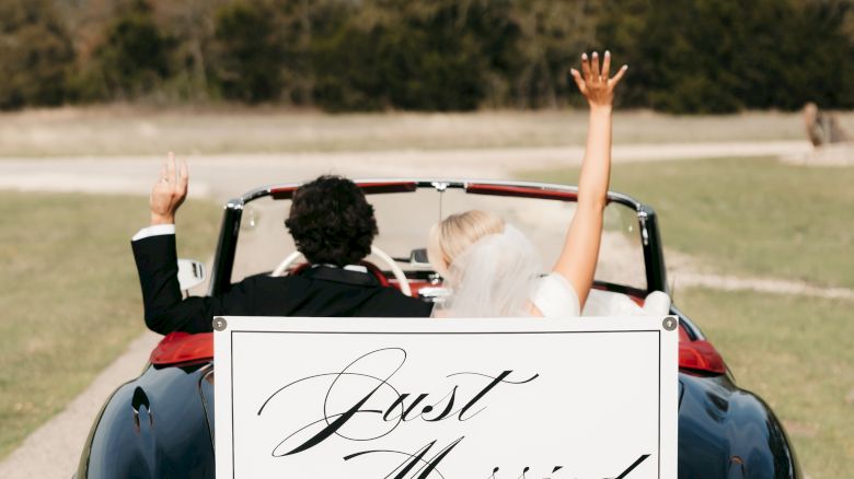 A couple waves from a classic blue convertible wedding car, with a &ldquo;Just Married&rdquo; sign for The Stewarts.