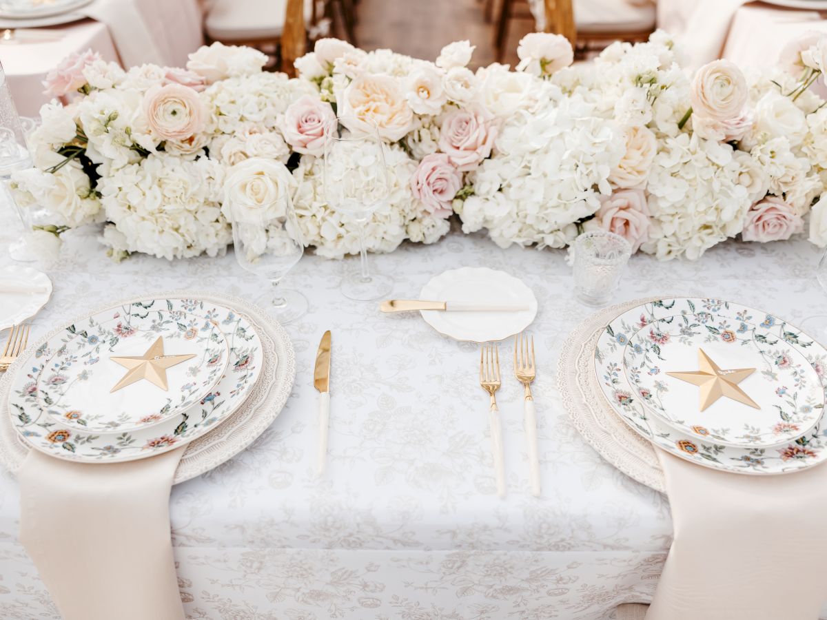 An elegant table setting with white and blush floral centerpiece, gold star plates, and gold cutlery on a pale patterned tablecloth.