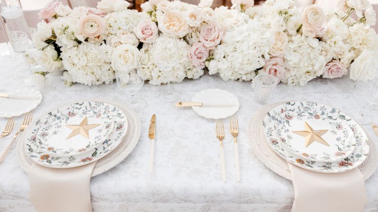 An elegant table setting with white and blush floral centerpiece, gold star plates, and gold cutlery on a pale patterned tablecloth.