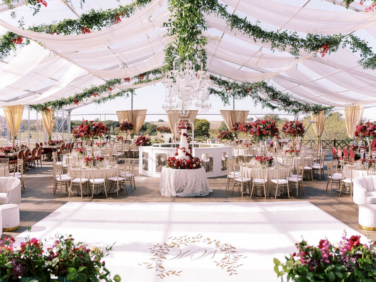 An elegant outdoor wedding reception under a white tent, decorated with lush greenery, red flowers, chandeliers, and neatly set tables.