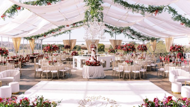 An elegant outdoor wedding reception under a white tent, decorated with lush greenery, red flowers, chandeliers, and neatly set tables.