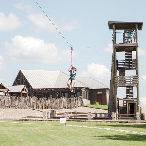 A person is ziplining from a tall tower, with buildings and a grassy area in the background on a bright, sunny day.