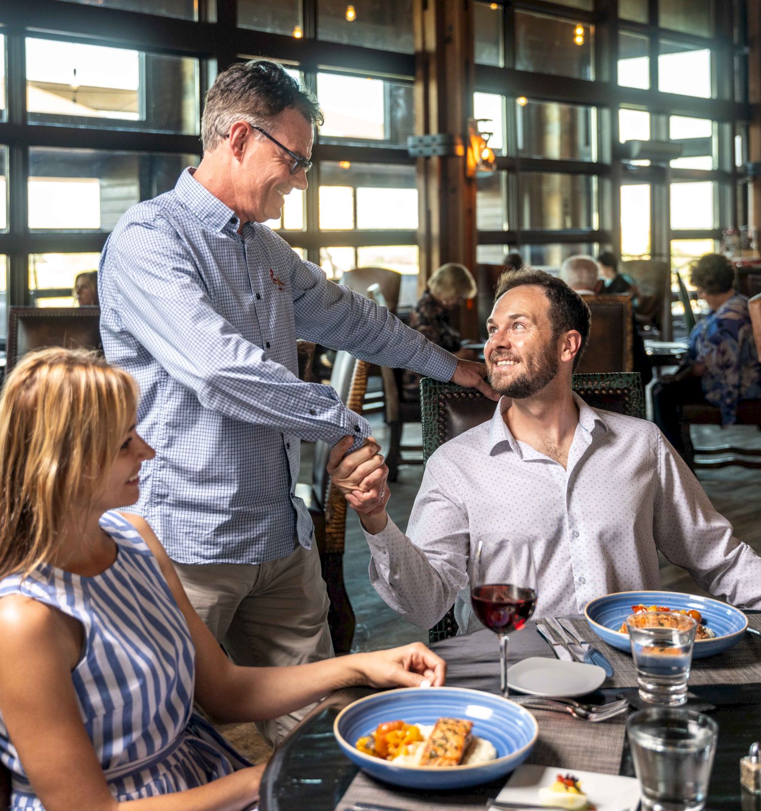 A man stands, shaking hands with a seated man at a restaurant table with food and wine, while a woman in a striped dress looks on, smiling.