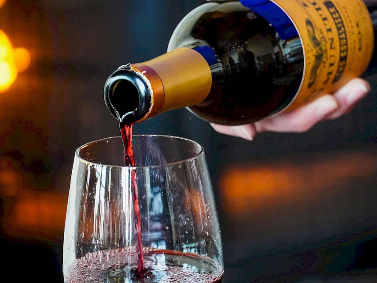 A hand pours red wine from a bottle into a glass, with a warm, blurry background lit by lights, creating an inviting atmosphere.