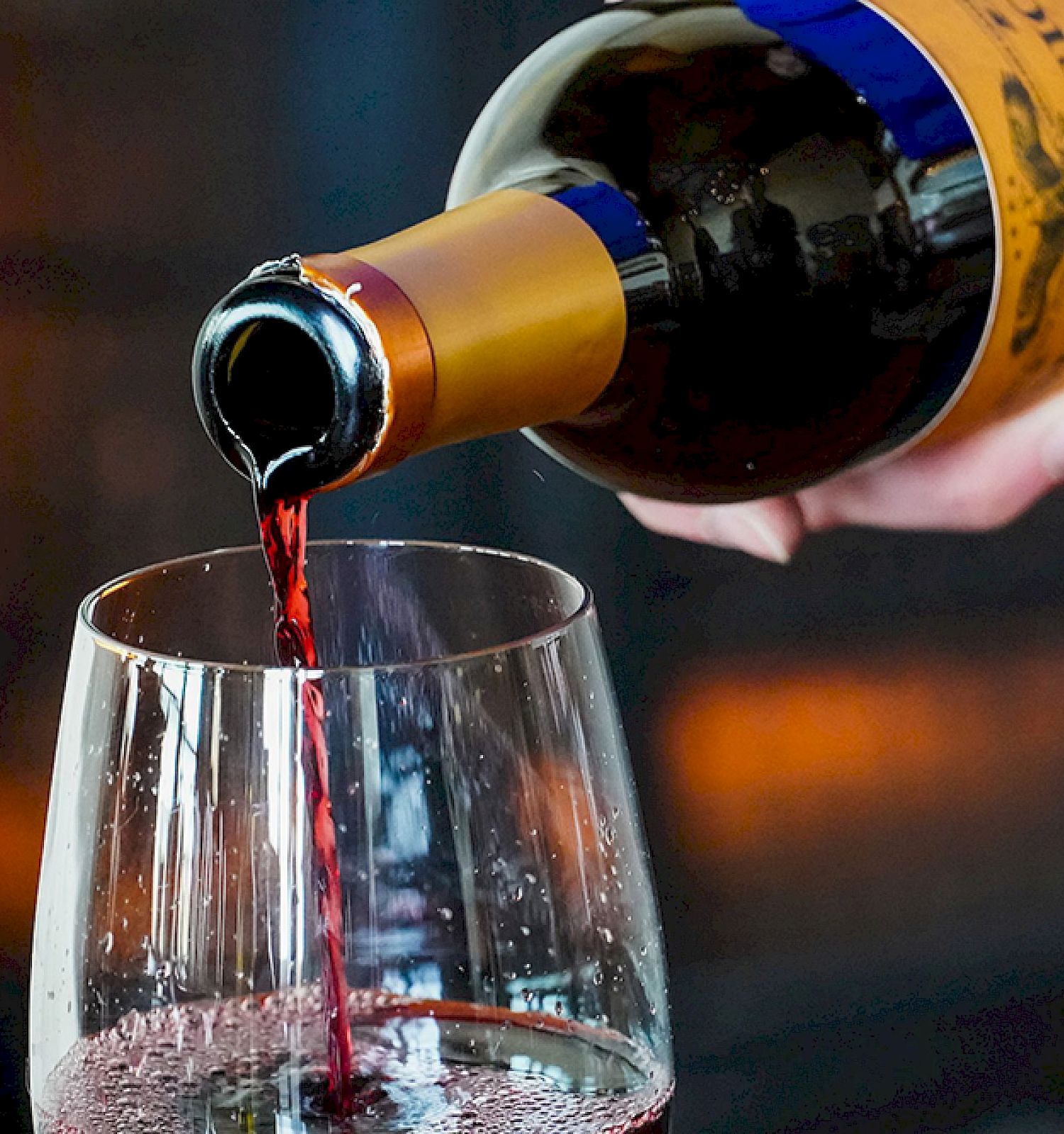 A hand pours red wine from a bottle into a glass, with a warm, blurry background lit by lights, creating an inviting atmosphere.