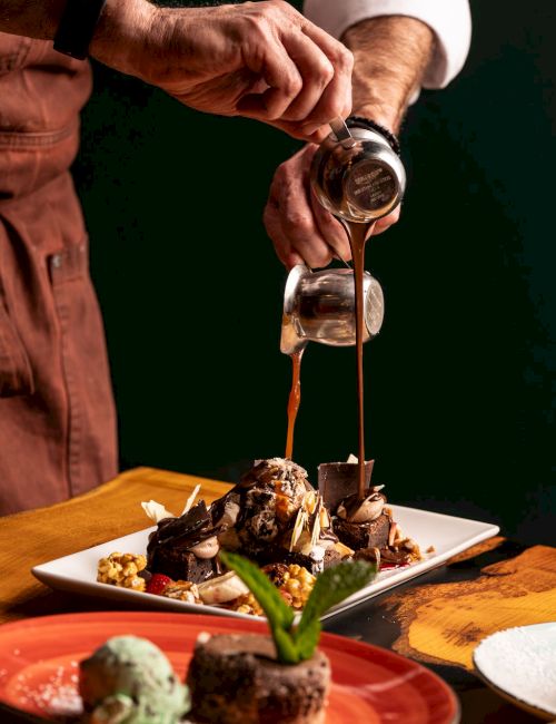 A chef pours rich caramel sauce from a small pitcher onto a plated chocolate dessert for a finishing flourish.