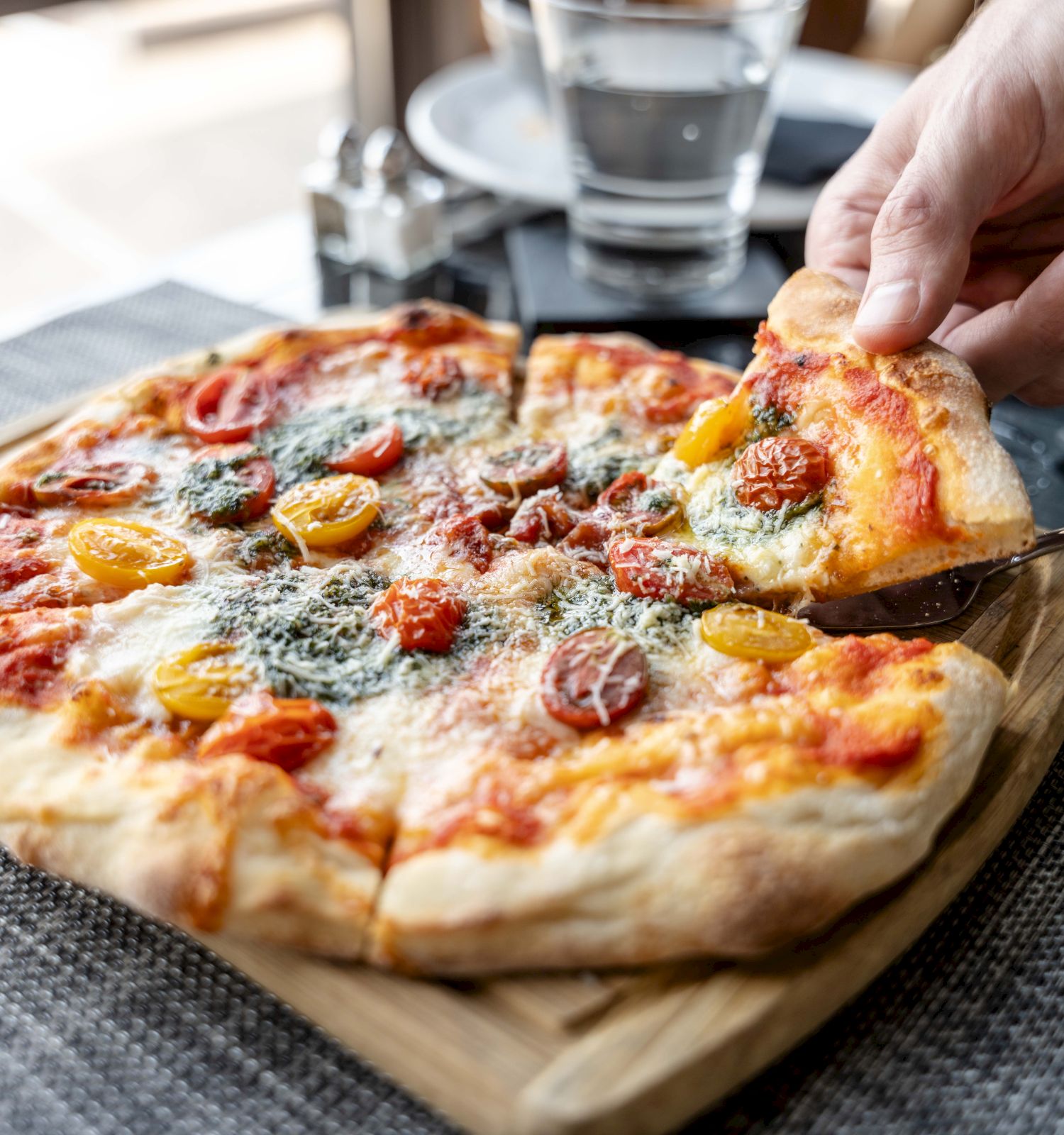 A person is lifting a slice of a pizza with toppings like cheese, tomatoes, and herbs, served on a wooden board.