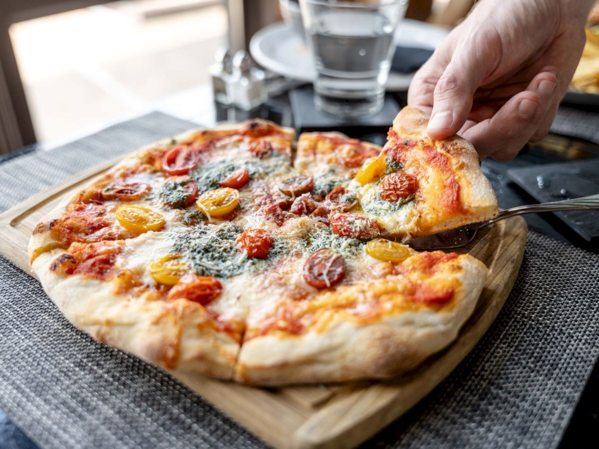 A hand is lifting a slice from a freshly baked pizza topped with tomatoes, basil, and cheese on a wooden board.