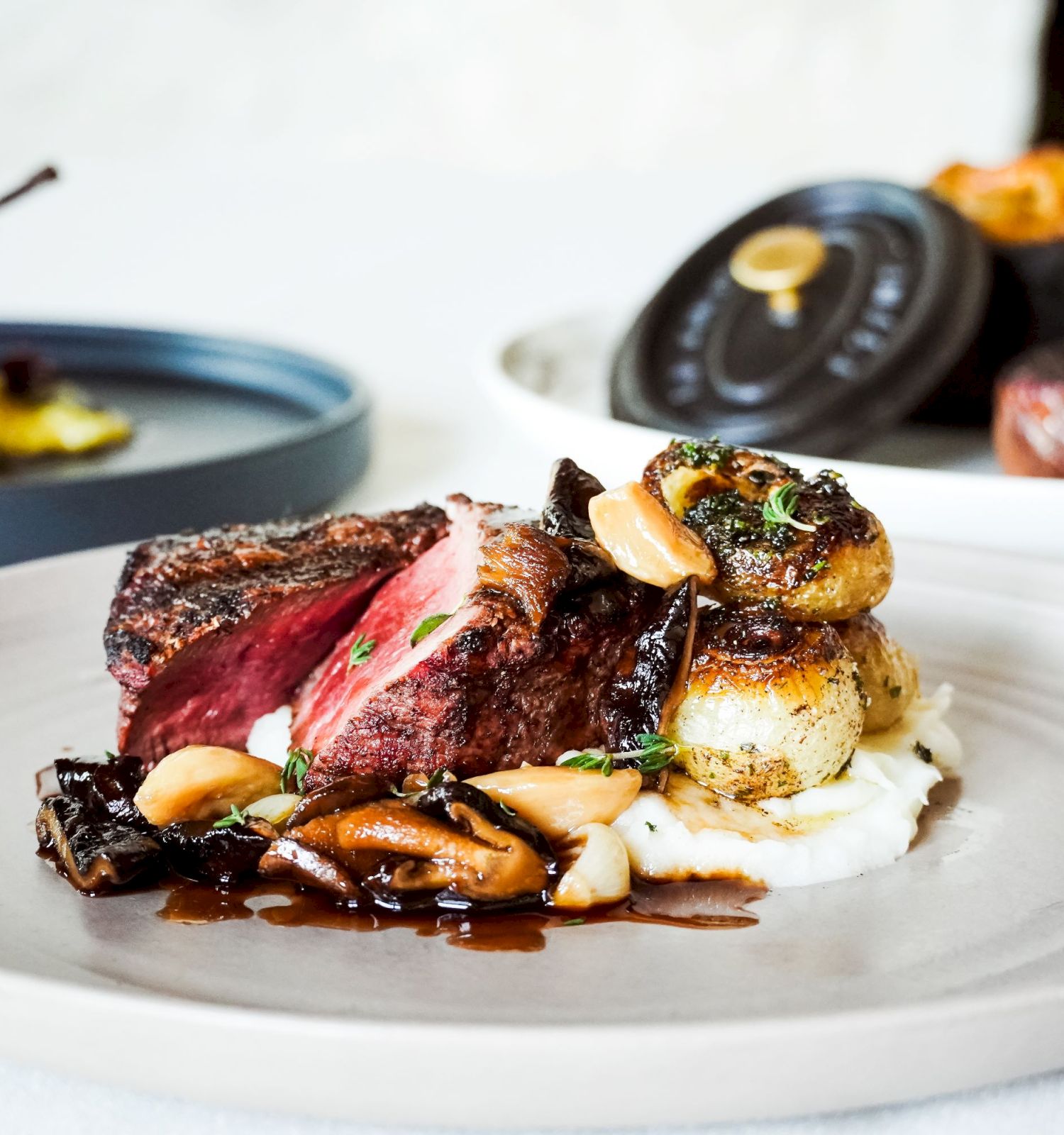 A gourmet dish features seared steak, roasted potatoes, and vegetables, elegantly plated. Other dishes are visible in the background.