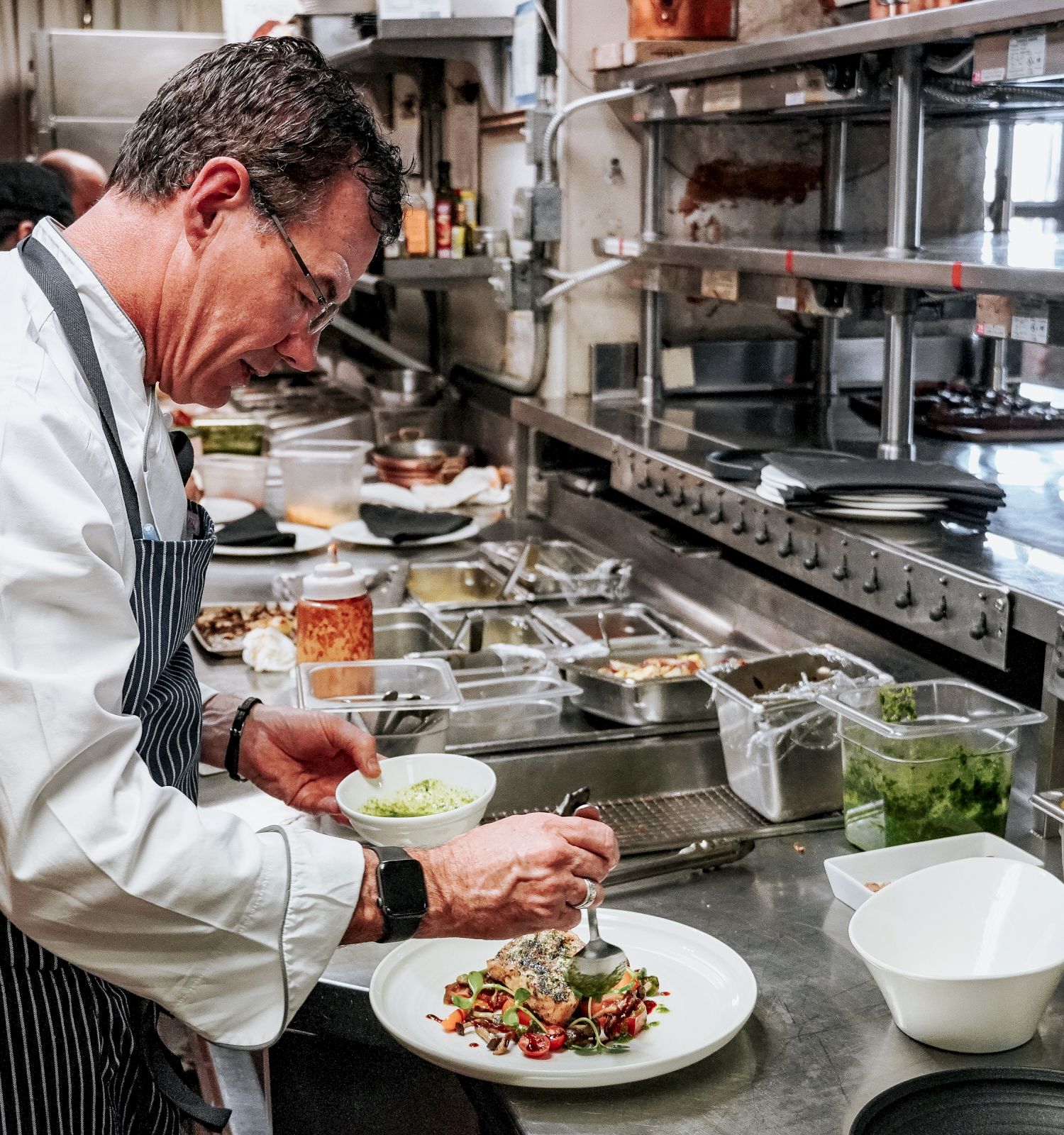 A chef is preparing a dish in a professional kitchen, garnishing a plate with a spoon, surrounded by various ingredients and utensils.