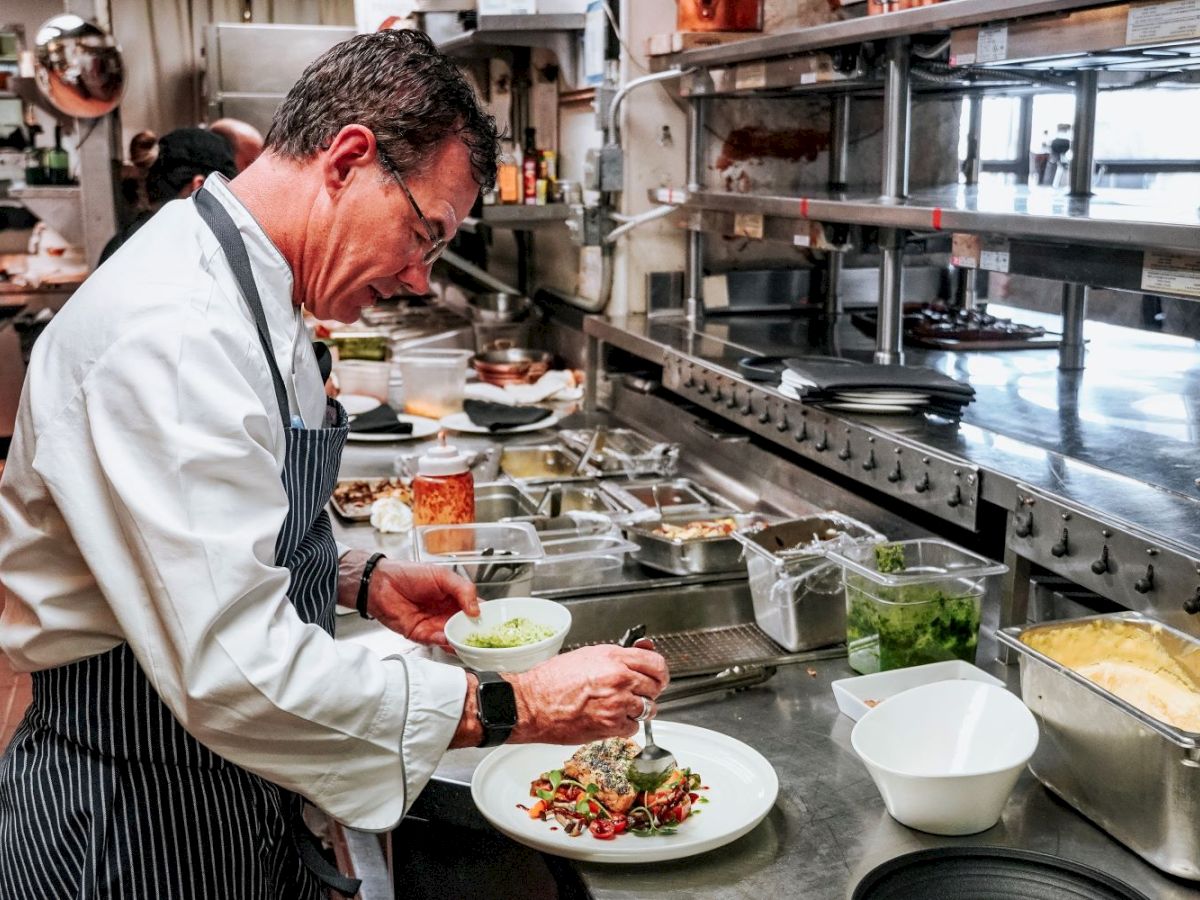 A chef in a professional kitchen is plating a dish, surrounded by various ingredients and cooking utensils, focusing intently on presentation.