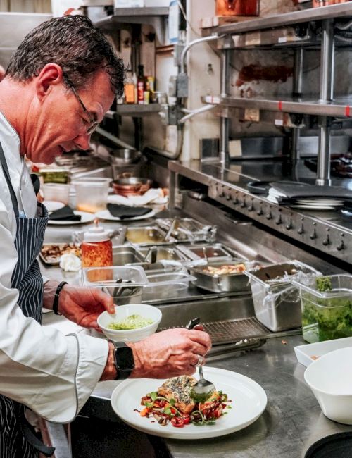 A chef in a professional kitchen is plating a dish, surrounded by various ingredients and cooking utensils, focusing intently on presentation.