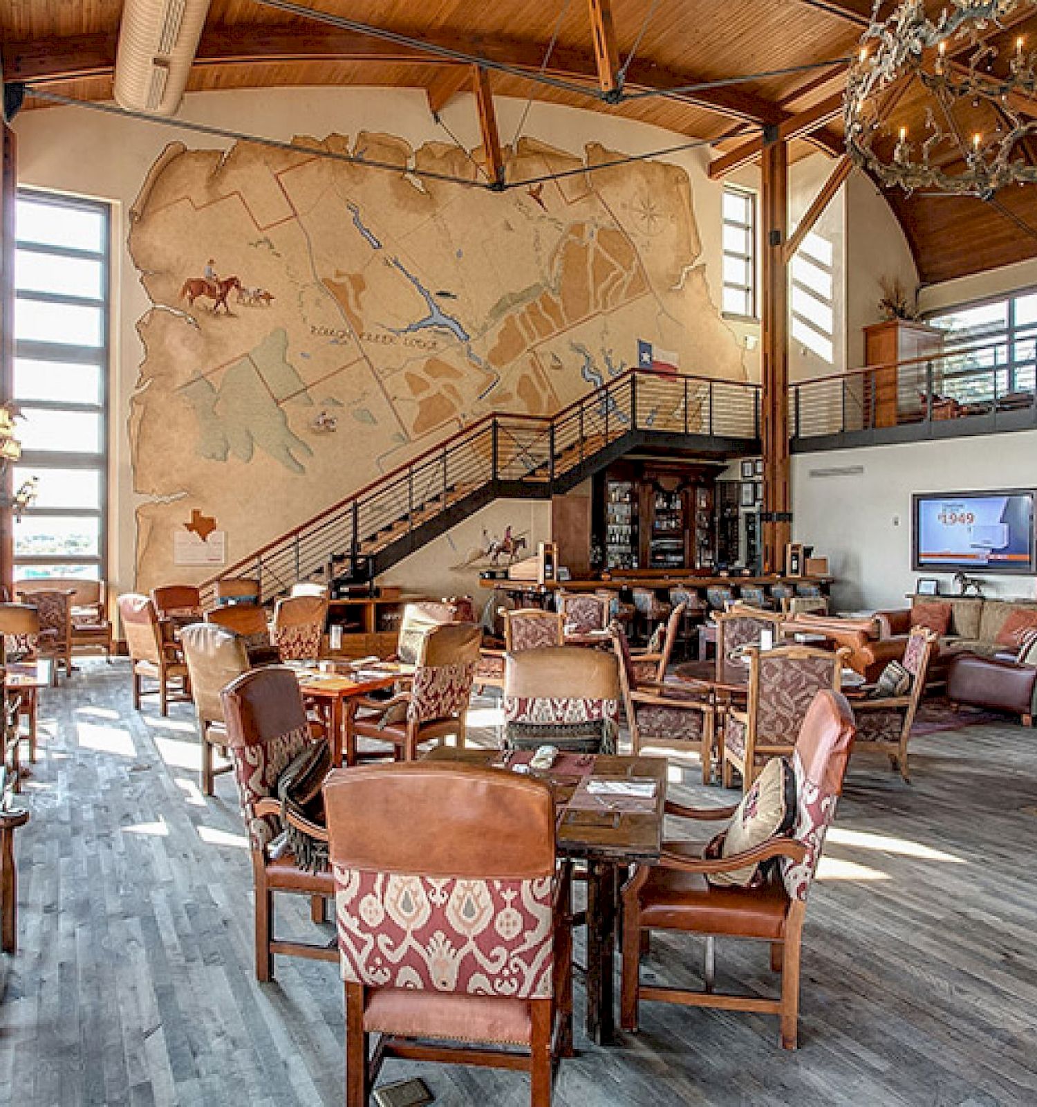 The image shows a spacious, rustic-themed restaurant with wooden furniture, large windows, a mural, a television, and a staircase.