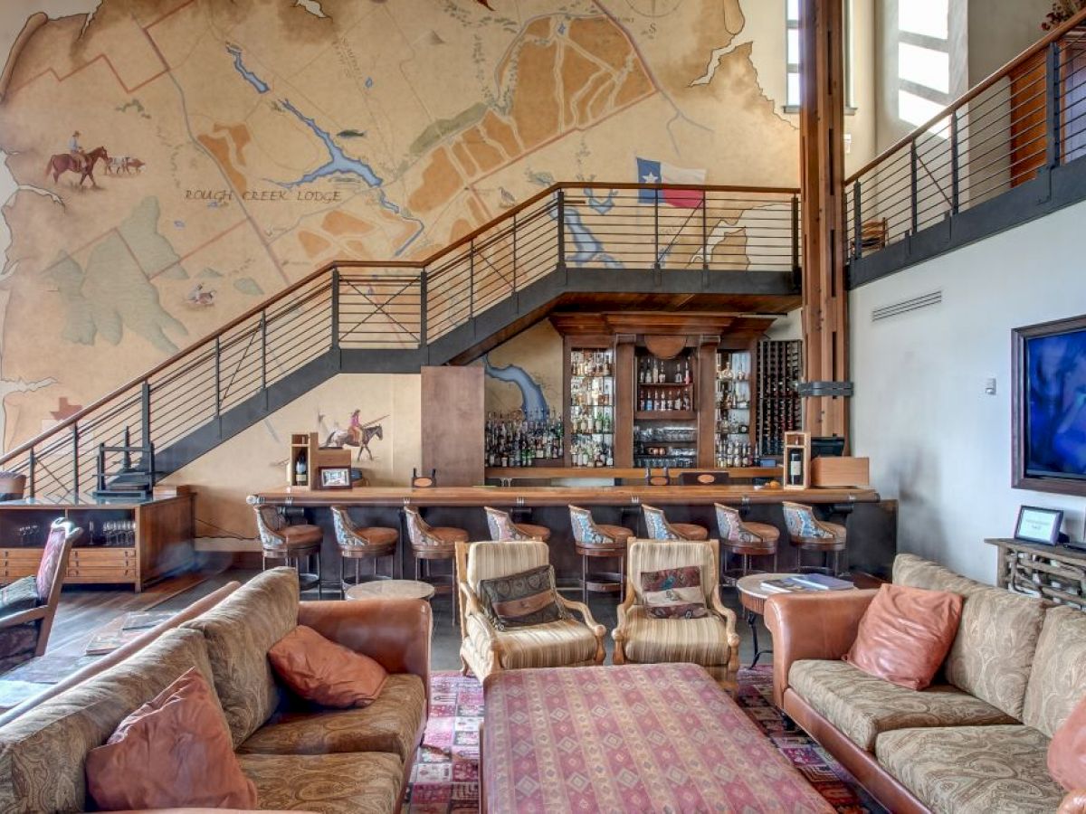 A cozy lounge with sofas, a bar area, a large mural map on the wall, and modern decor. There's a staircase and a flat-screen TV.