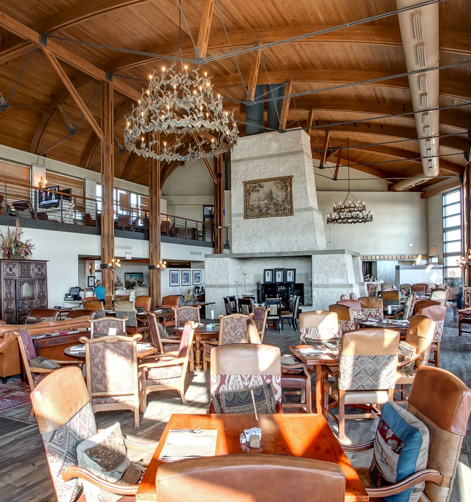 A spacious restaurant with wooden beams, a large stone fireplace, chandeliers, and various seating arrangements under a high ceiling.