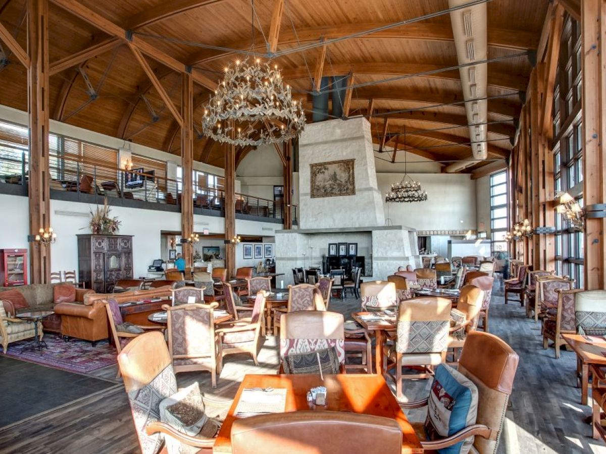 A spacious, rustic lodge-style interior with wooden beams, chandeliers, and a large stone fireplace, featuring ample seating and natural light.