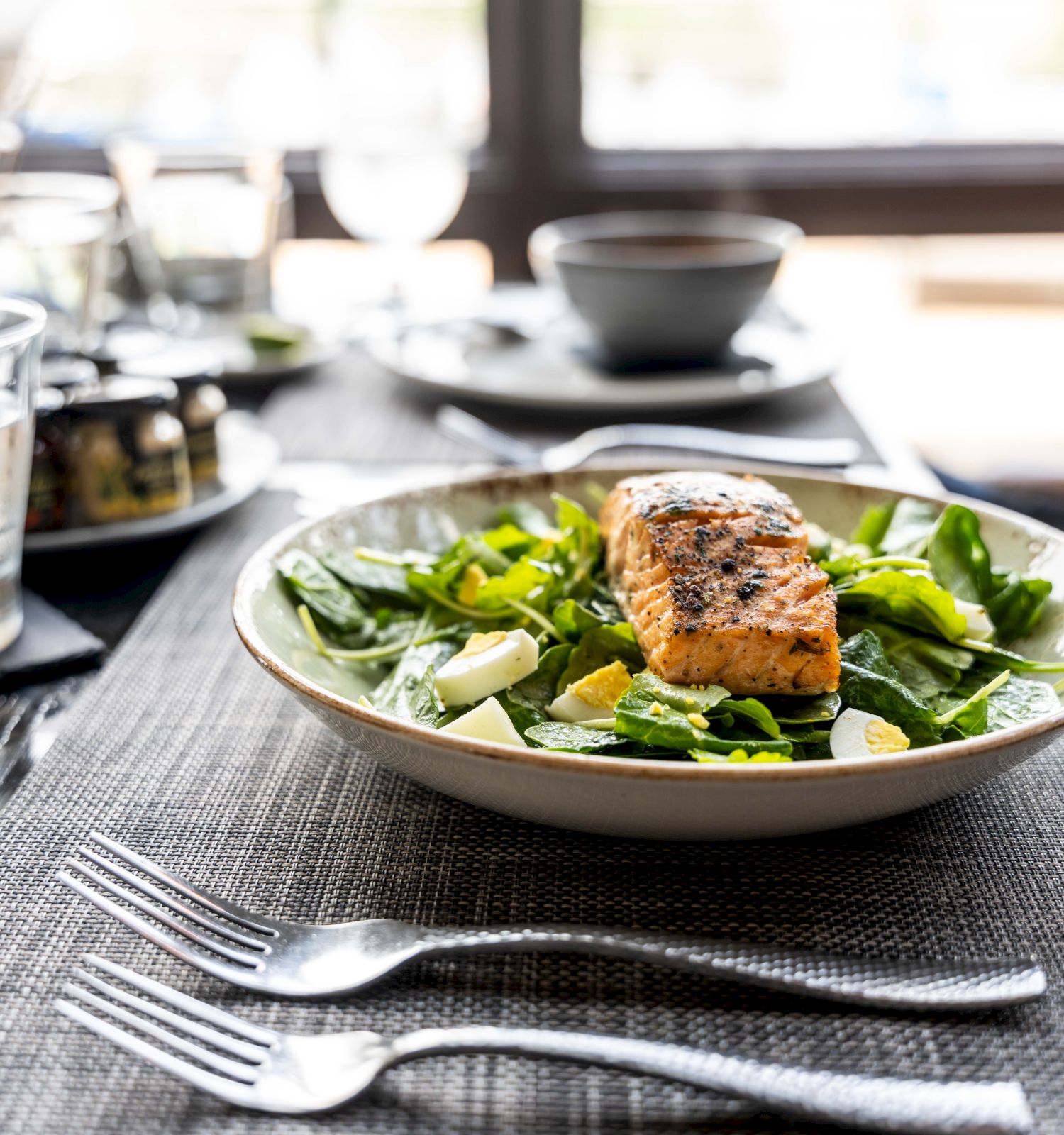 A plate of grilled salmon on a bed of greens, lemon wedges, and other dishes in the background on a dining table.