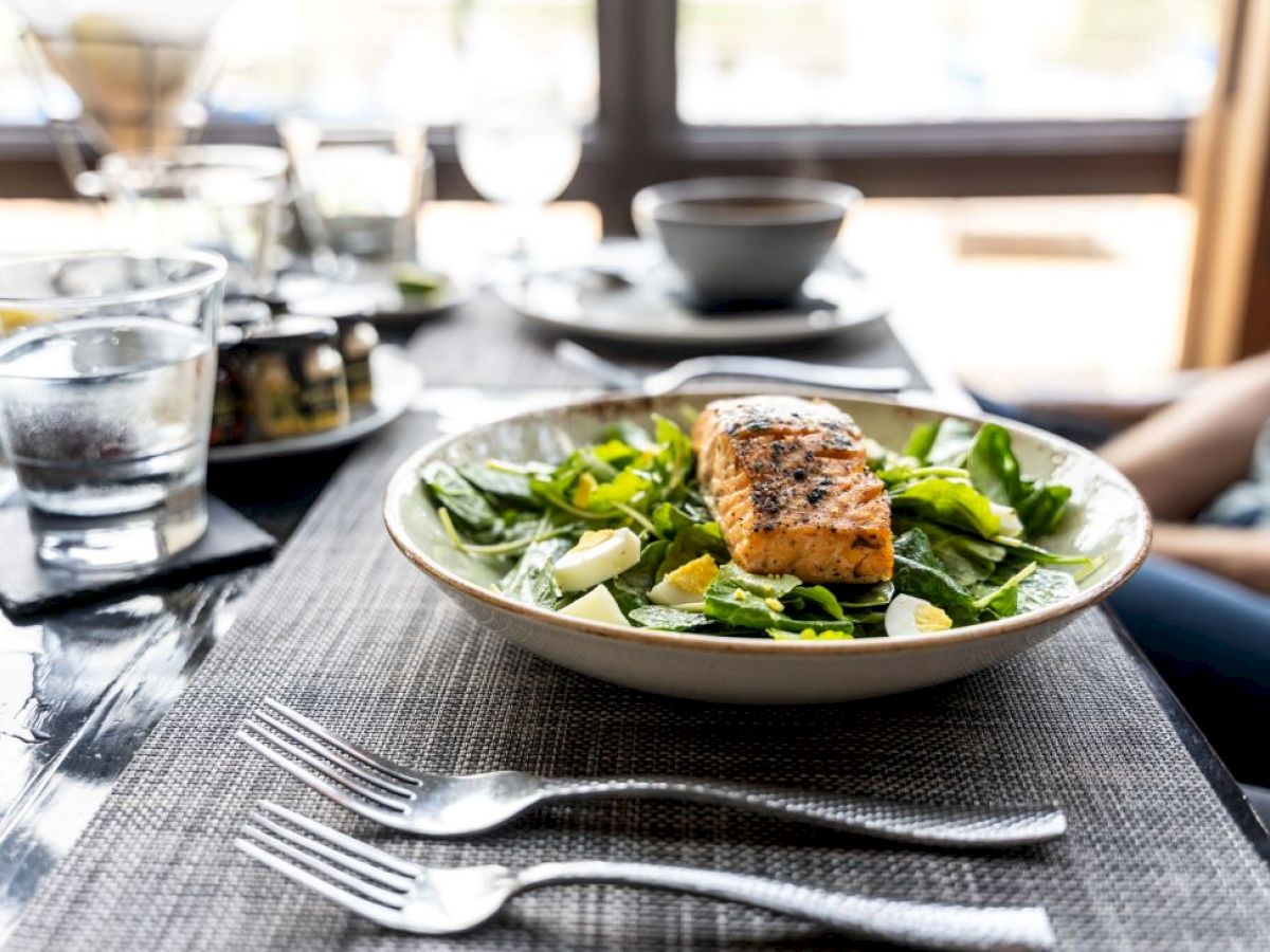 A plate of grilled salmon on spinach and greens, surrounded by cutlery, water glasses, and other dishes on a dining table.