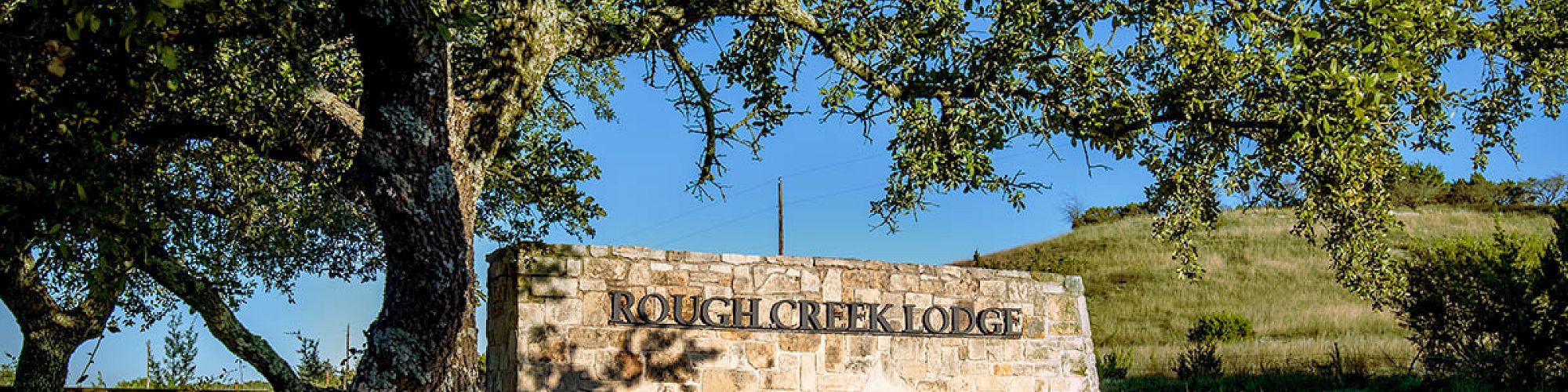 The image shows a stone sign that reads "Rough Creek Lodge," situated in a grassy area with trees and a hill in the background, under a clear blue sky.