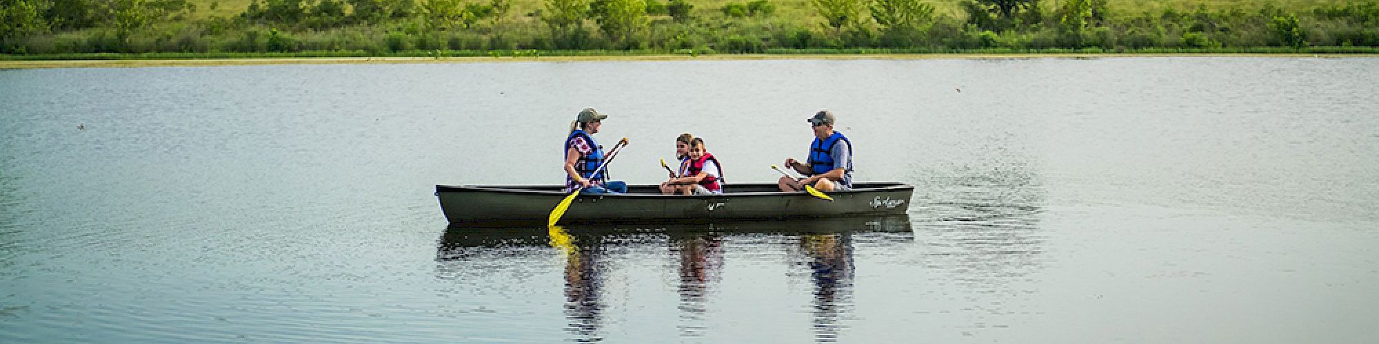 Three people are in a canoe on a calm body of water, with grassy fields and trees in the background.