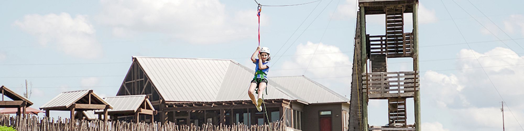 A person is zip-lining from a tall wooden tower in front of rustic buildings under a partly cloudy sky.