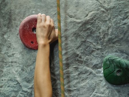 The image shows a person’s hand gripping a red climbing hold on an indoor rock climbing wall, with a green hold nearby.