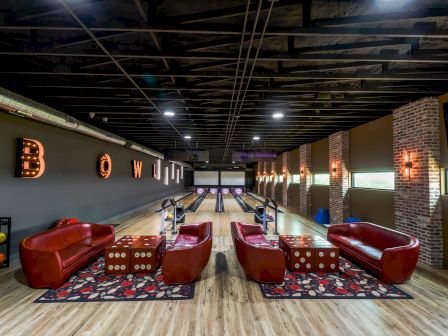 A stylish bowling alley with red sofas, dice-themed tables, colorful bowling balls, and illuminated "BOWL" letters on the wall.