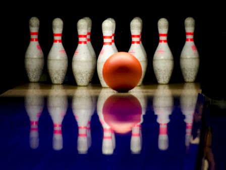 A bowling ball is about to strike several bowling pins, lined up in a classic formation on a reflective surface in a bowling alley.