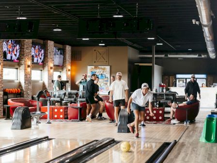People are bowling in a modern alley with brick walls, TV screens, and lounge seating, creating a fun atmosphere.