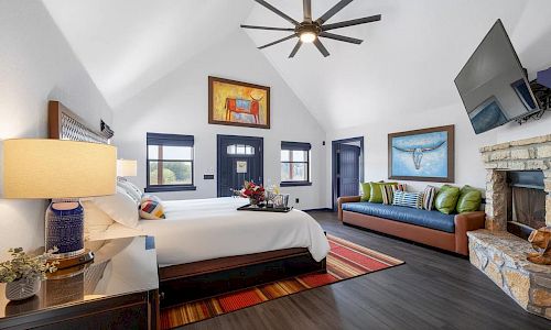 A bright, spacious hotel room with a high, vaulted ceiling, a large bed, a blue-trimmed doorway, a sofa with green cushions, a stone fireplace, wall-mounted TV, and contemporary decor.