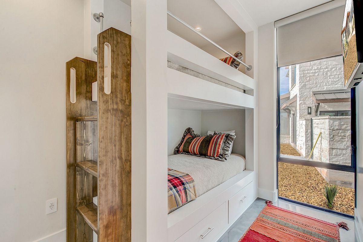 A modern bedroom with bunk beds, colorful pillows, a wooden ladder, and a large window, offering a view outside.