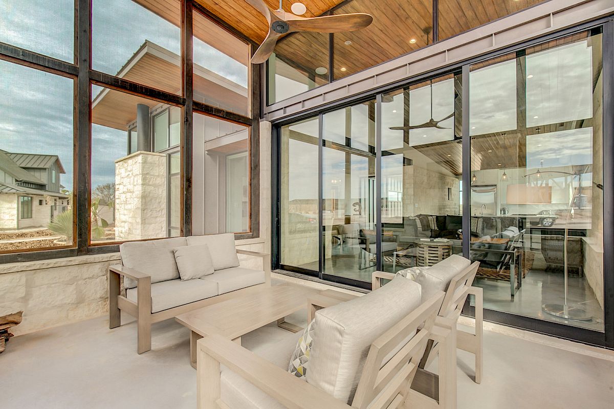 The image shows a modern patio with beige furniture, large glass windows, and a view into a contemporary living room.