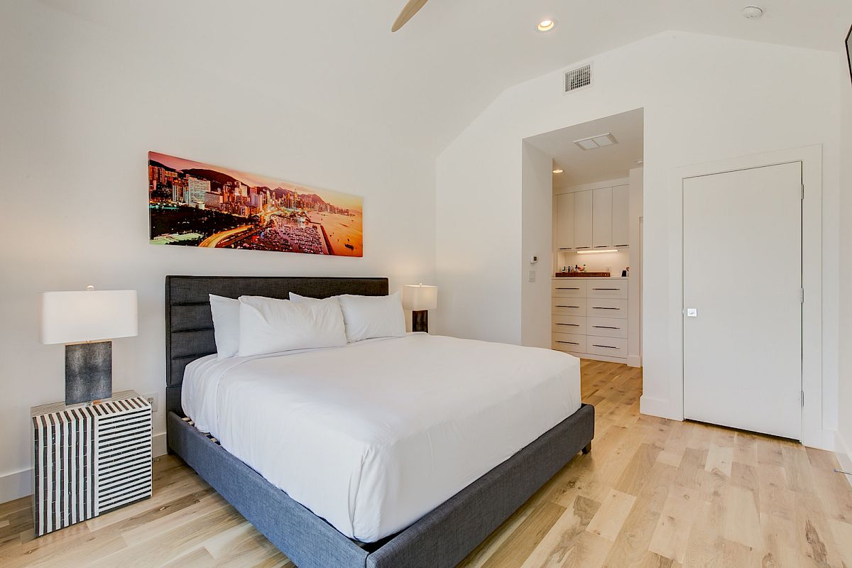 A modern bedroom with a large bed, nightstand, cityscape artwork, and clean, minimalist design on a wooden floor.