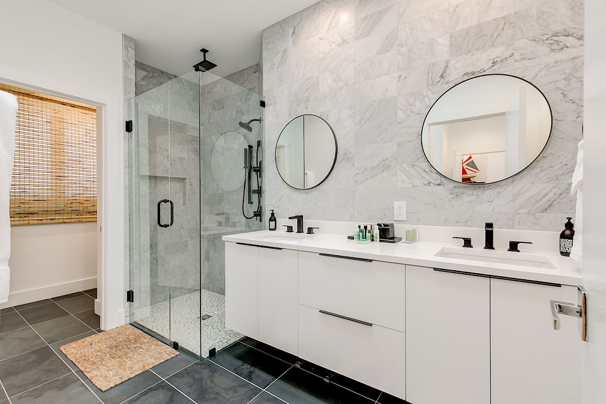Modern bathroom with dual sinks, round mirrors, a glass shower, and marble walls. Black fixtures and a neutral palette complete the look.