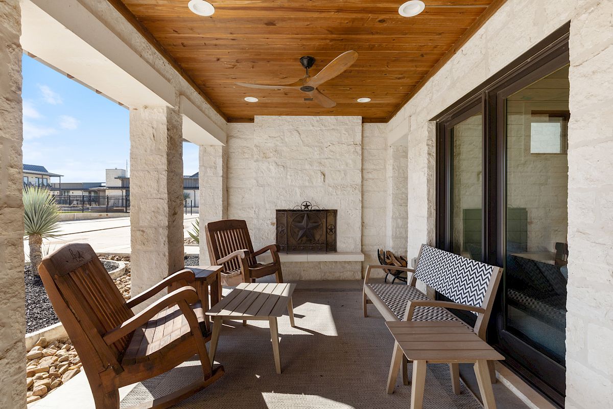 A cozy patio with a wooden ceiling, fan, fireplace, rocking chairs, and cushioned bench, overlooking a sunny outdoor area.