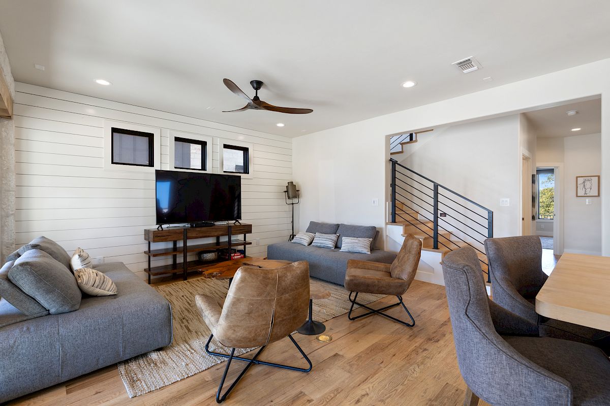 A modern living room with a TV, gray sofas, brown chairs, ceiling fan, shiplap walls, and stairs leading up.