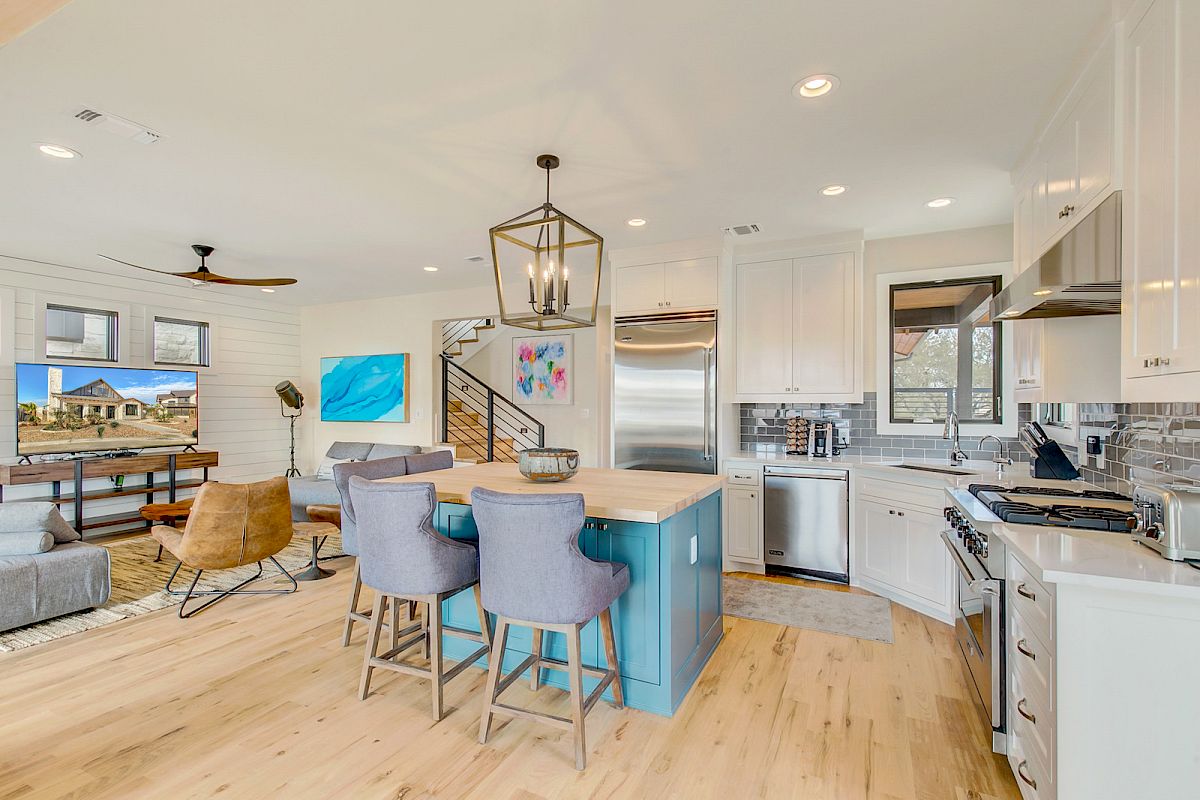 A modern open-plan kitchen and living area with a central island, dining area, artwork on walls, and wooden floors.