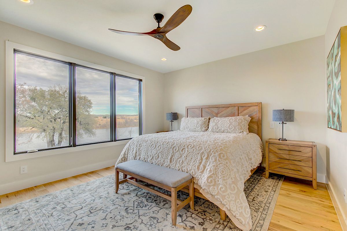A bedroom with a bed, wooden furniture, ceiling fan, large window view, and decorative rug.