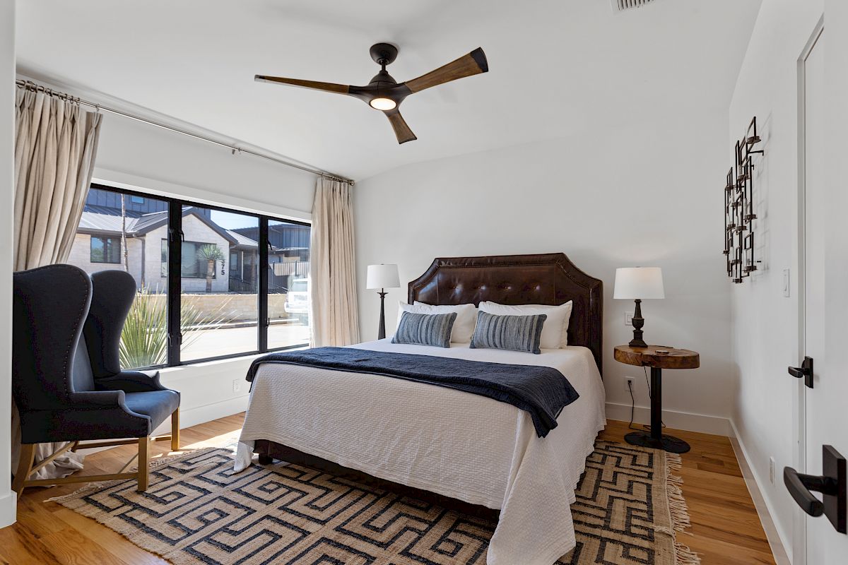 A bedroom featuring a bed with a headboard, a wingback chair, lamps on nightstands, a geometric rug, and a ceiling fan.