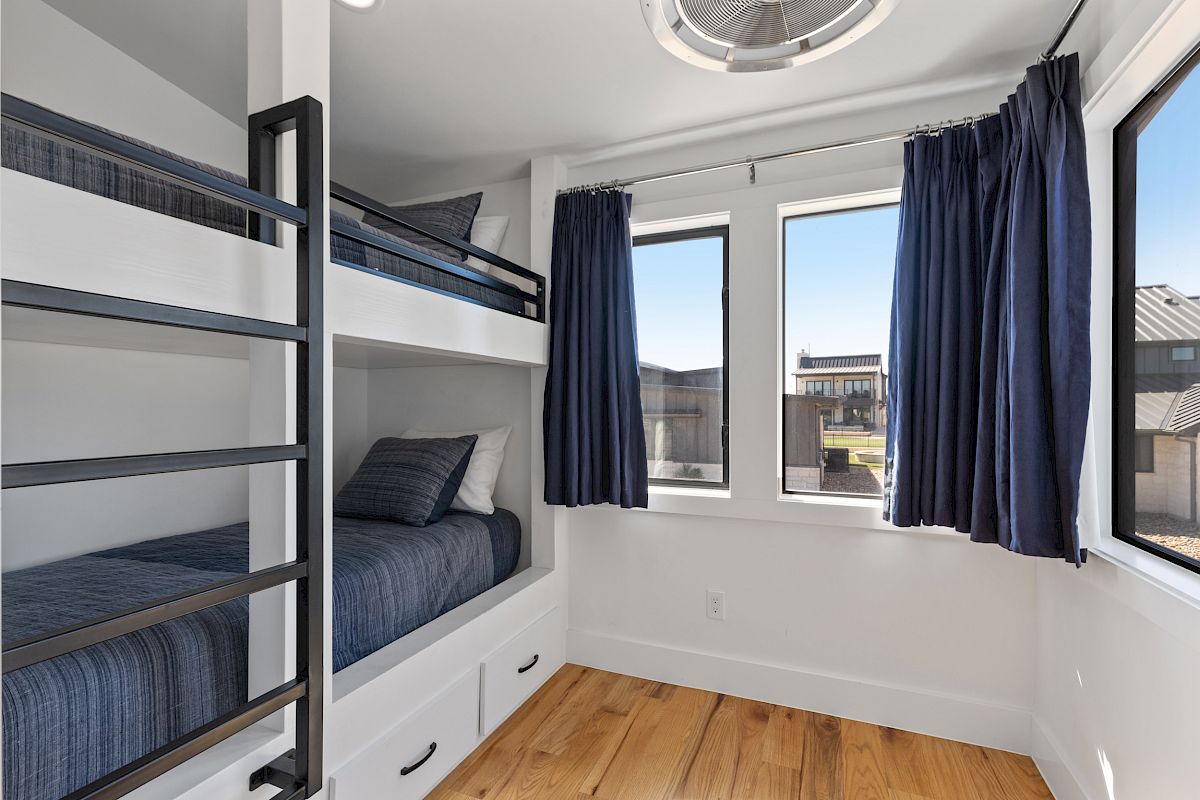 The image shows a small bedroom with a bunk bed, blue bedding, curtains, and sunlight streaming through the windows.