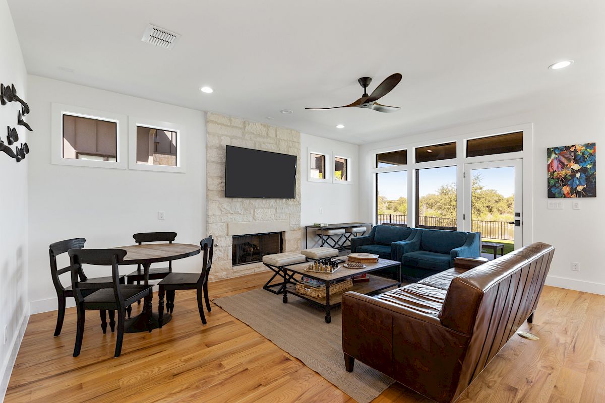 A modern living room with a sofa, chairs, a round dining table, a wall-mounted TV, artwork, and large windows with a scenic view.