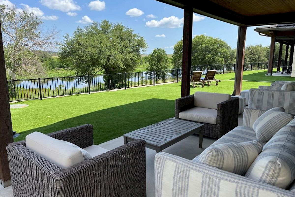 A covered patio with cushioned wicker furniture overlooks a lush lawn, trees, and a river. Two loungers are in the background.