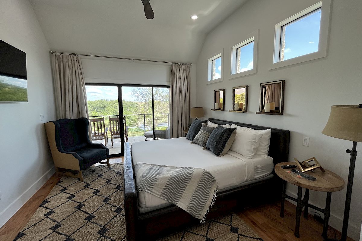 A modern bedroom with a bed, armchair, and side tables. Large windows and glass doors offer a view outside, with stylish decor throughout.