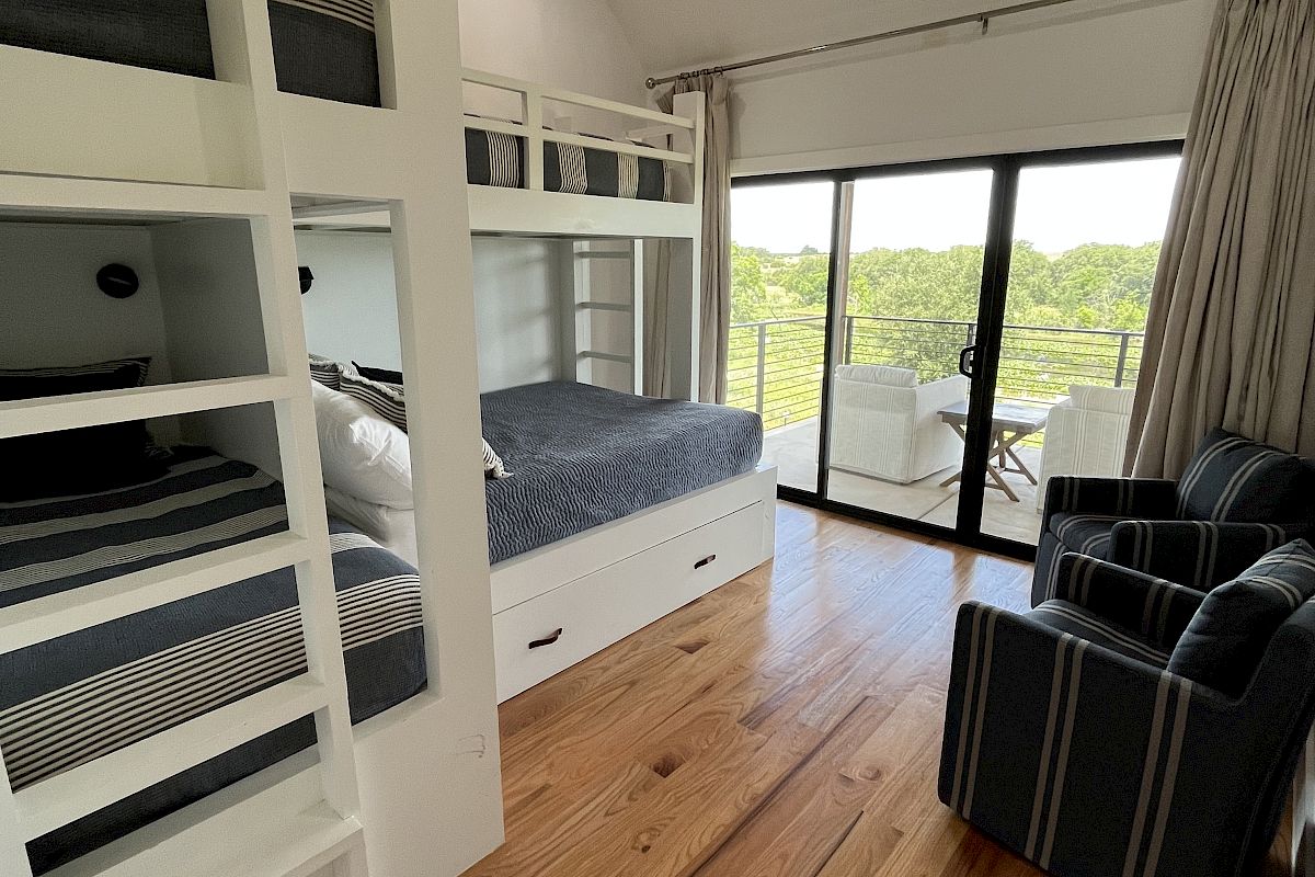 A bright room with bunk beds, striped chairs, and large glass doors opening to a balcony with a scenic view.