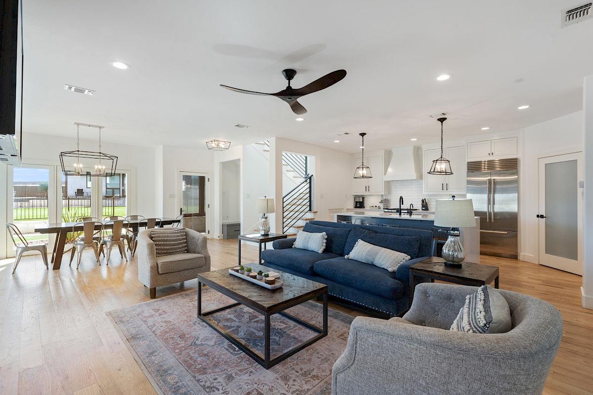 The image shows a modern open-concept living space with a blue sofa, chairs, dining area, and kitchen featuring pendant lights and stainless appliances.