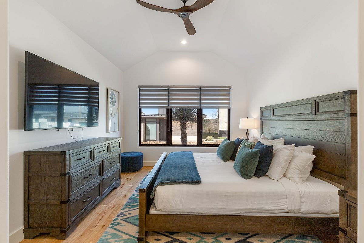 A cozy bedroom with a large bed, wooden furniture, a TV, and a ceiling fan, illuminated by natural light through a large window.