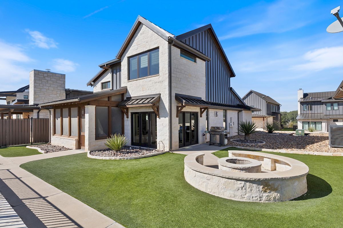 A modern house with stone and metal exterior, landscaped yard, patio with grill, and fire pit in a residential neighborhood.