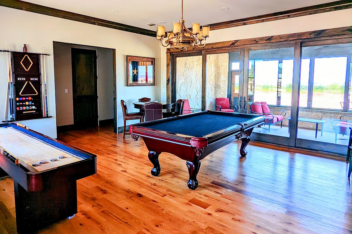 A game room with a pool table, shuffleboard, large windows, and a chandelier, featuring wooden floors and outdoor views.