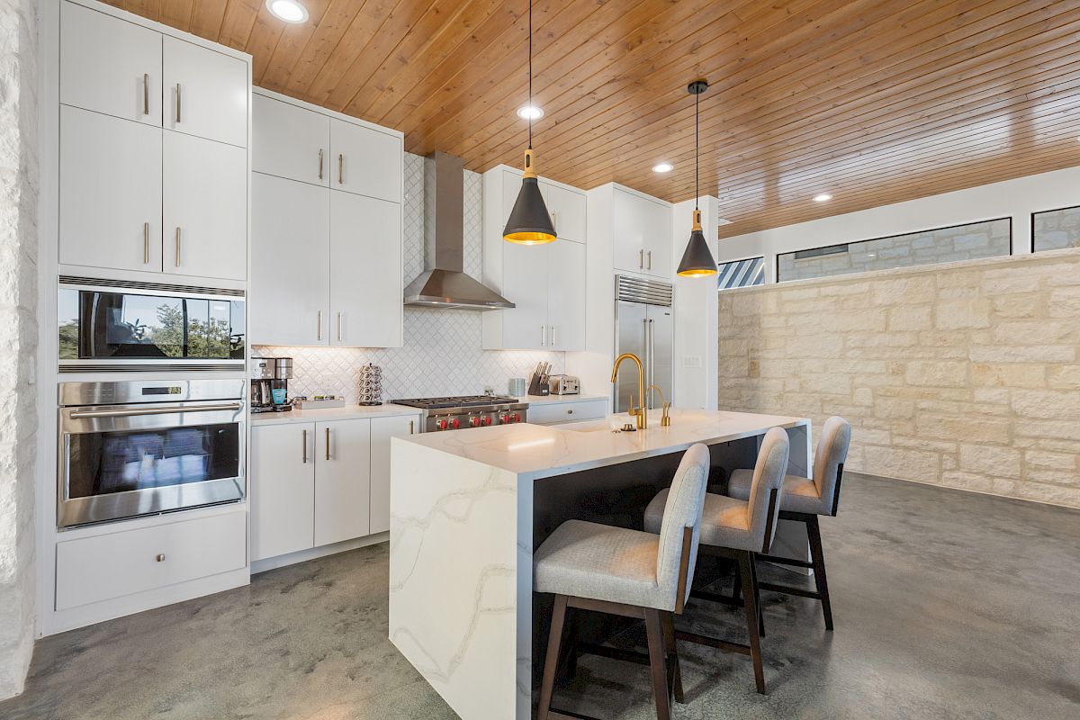 Modern kitchen with white cabinets, island with seating, pendant lights, and stainless steel appliances, featuring a wooden ceiling.