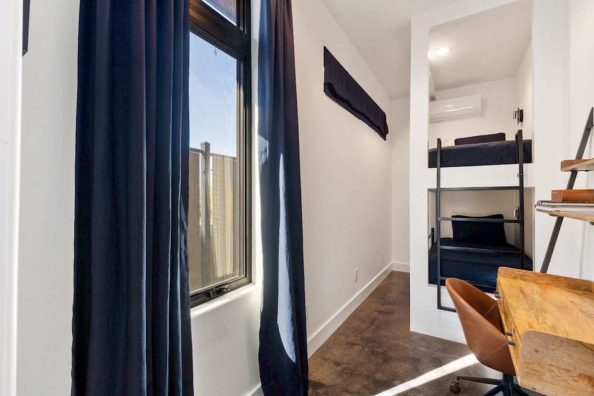 A small room featuring a window, black curtains, bunk beds, a wooden desk, and an office chair; the decor is simple and modern.
